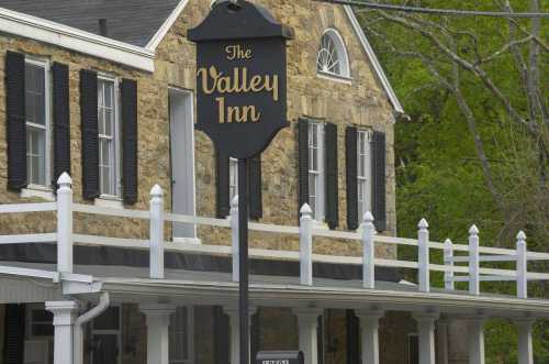 Sign for "The Valley Inn" in front of a stone building with black shutters and a white railing. Green trees in the background.