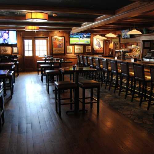 A cozy bar interior with wooden floors, high tables, and a long counter, featuring TVs and framed artwork on the walls.