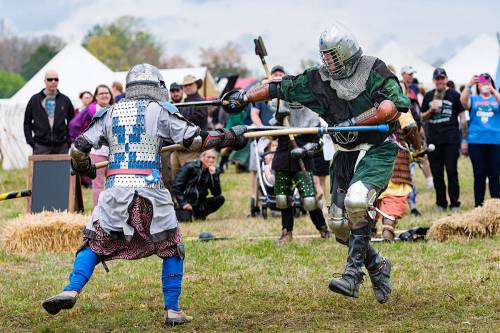 Two armored knights engage in a mock battle with wooden weapons in a grassy field, surrounded by spectators.