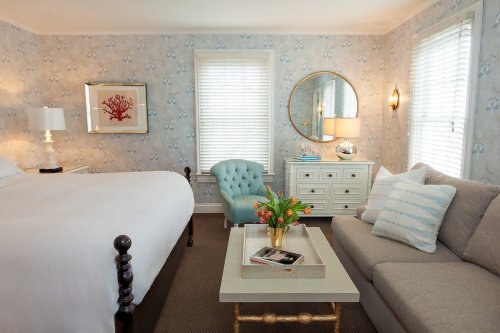 Cozy bedroom with a bed, blue accent chair, coffee table, and floral wallpaper, featuring natural light from windows.