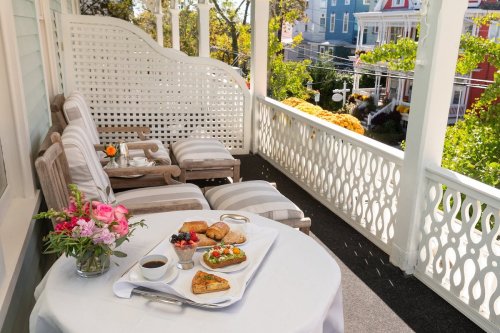 A cozy porch with a table set for breakfast, featuring flowers, pastries, and coffee, overlooking colorful houses.