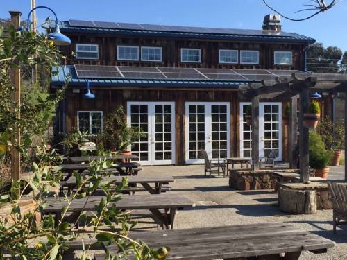 A rustic building with solar panels, surrounded by outdoor seating and greenery under a clear blue sky.
