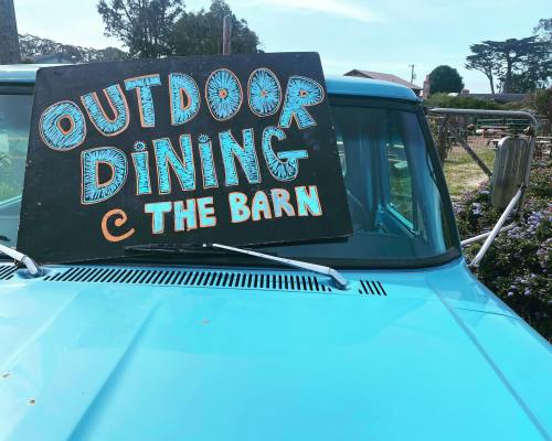 A turquoise truck with a sign reading "Outdoor Dining @ The Barn" displayed on the windshield.
