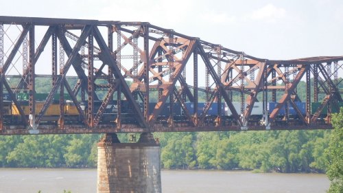 A rusty train bridge spans a river, with a freight train carrying colorful containers crossing above.