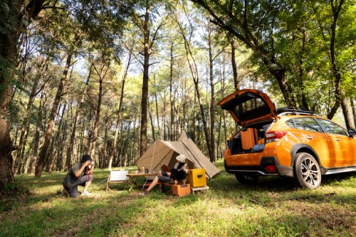 A person photographs a campsite with a tent, orange car, and camping gear surrounded by tall trees in a forest.