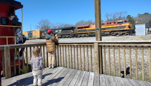Two children watch a passing train from a wooden platform, with a red train car in the background under a clear blue sky.