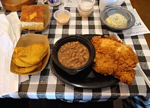 A plate with fried chicken, pinto beans, cornbread, and fried tortillas on a checkered tablecloth.