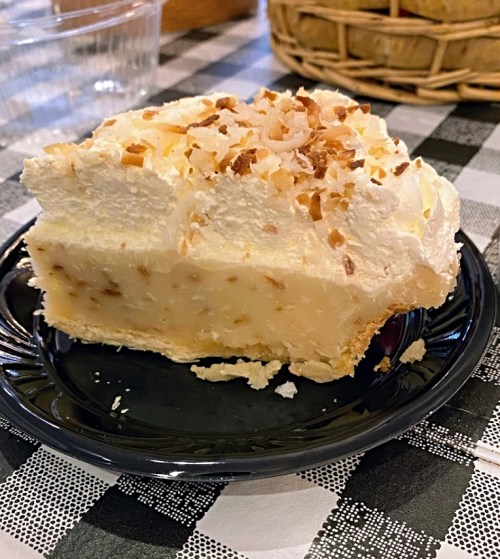 A slice of creamy coconut pie topped with whipped cream and toasted coconut flakes on a black plate.