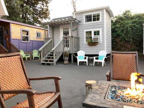 Cozy outdoor scene with a fire pit, chairs, and a small house surrounded by greenery.