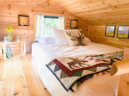 Cozy wooden cabin interior featuring a bed with a quilt, decorative pillows, and natural light from a window.