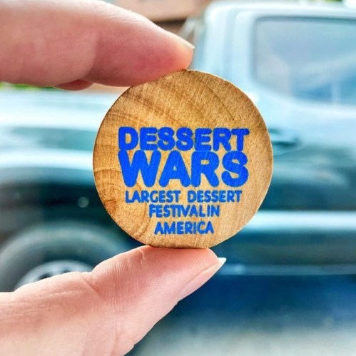 A hand holding a wooden token that reads "DESSERT WARS LARGEST DESSERT FESTIVAL IN AMERICA."