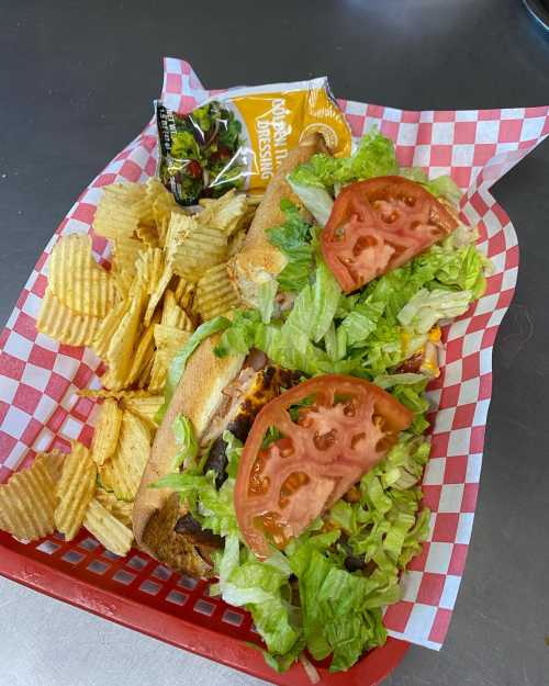 A basket with a sandwich loaded with lettuce and tomato, served with potato chips and a packet of dressing.