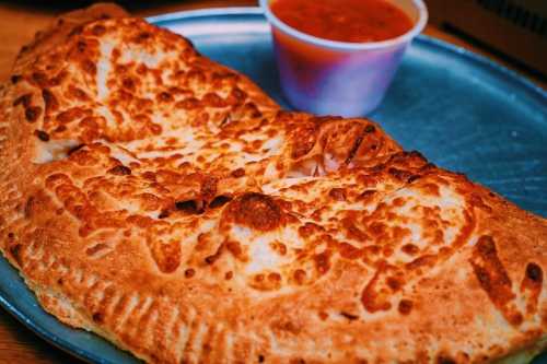 A golden-brown calzone on a blue plate, accompanied by a small cup of marinara sauce.