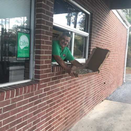 A smiling man in a green shirt hands a pizza out of a window at a brick building.