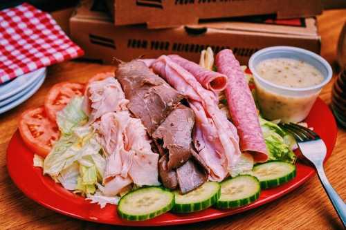 A colorful plate of assorted deli meats, lettuce, tomatoes, cucumbers, and a side of dressing on a wooden table.