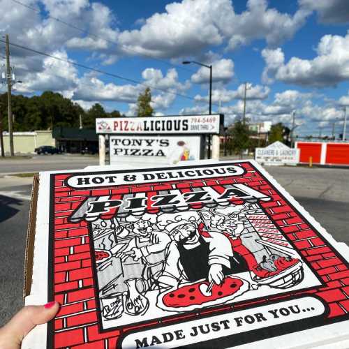 A pizza box with a cartoon chef and pizza graphics in front of a pizzeria under a blue sky with fluffy clouds.