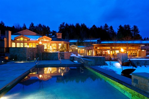 A serene evening view of a modern building with large windows, reflecting in a pool, surrounded by snow and trees.