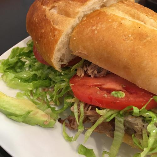 A close-up of a sandwich with shredded meat, lettuce, and tomato on a toasted baguette, served on a white plate.