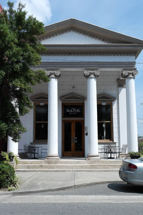 Historic bank building with tall columns, large wooden door, and outdoor seating, surrounded by greenery and parked cars.