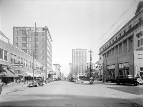 Black and white photo of a city street lined with buildings, shops, and vintage cars from the early 20th century.