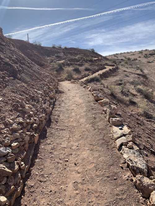 A winding dirt path lined with stones, surrounded by dry terrain and sparse vegetation under a clear blue sky.