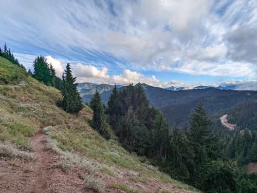 A scenic mountain landscape with lush green trees, a winding path, and a cloudy blue sky in the background.