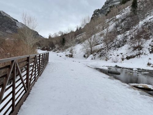Snow-covered path alongside a river, with a wooden bridge and mountains in the background under a cloudy sky.