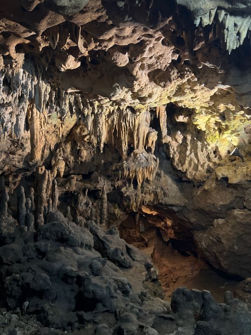 A dimly lit cave interior featuring stalactites and stalagmites, with textured rock formations and shadows.