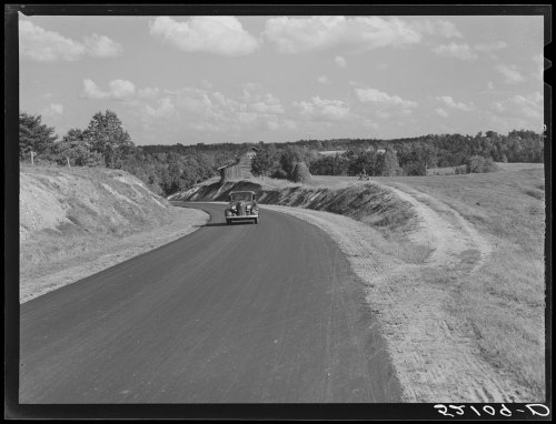 A vintage car drives along a winding road surrounded by fields and trees under a cloudy sky.