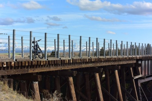 A bicycle rests on a wooden bridge with a railing, overlooking a scenic landscape and distant snow-capped mountains.