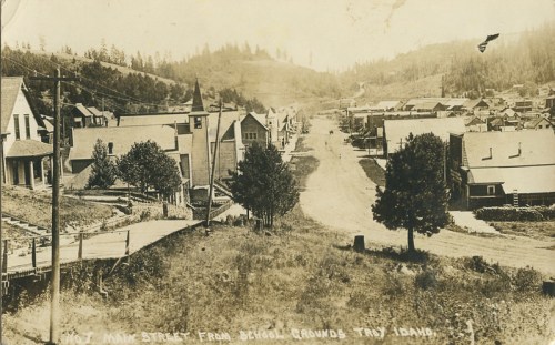 Historic view of Main Street from school grounds in Troy, Idaho, featuring wooden buildings and a dirt road.