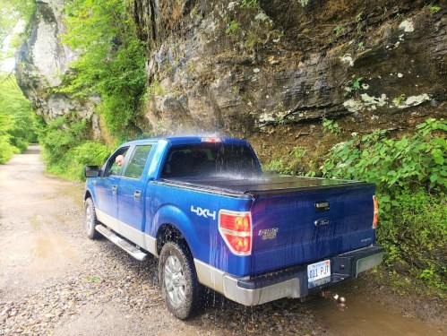 A blue Ford truck parked on a gravel road near a rocky cliff, with water cascading down the rocks.