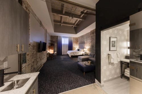 A stylish hotel room featuring exposed brick walls, a bed, seating area, and modern bathroom fixtures.