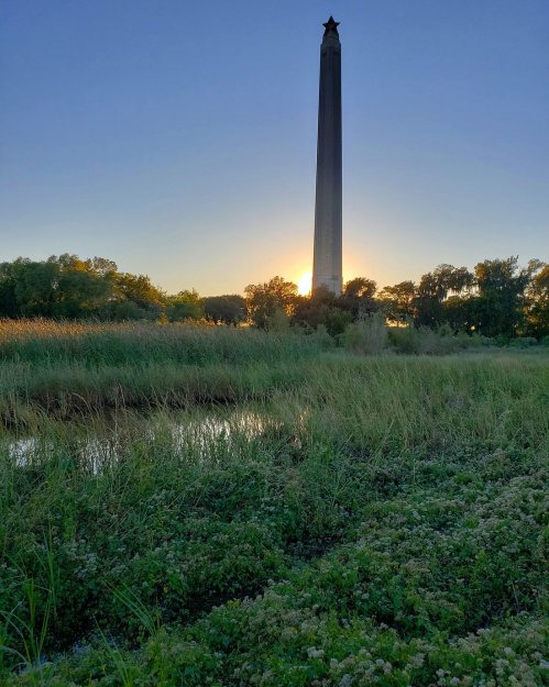 A tall monument stands against a sunset, surrounded by lush greenery and a calm waterway.