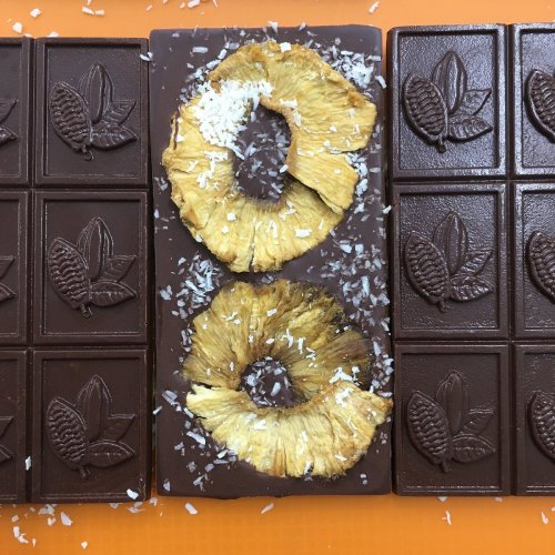 A chocolate bar topped with dried pineapple slices and sprinkled with shredded coconut on an orange background.