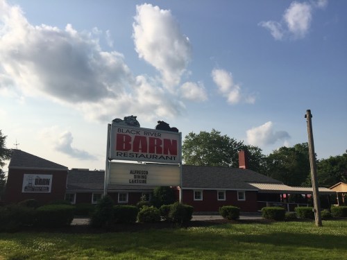 Sign for Black River Barn Restaurant with a blue sky and clouds in the background, surrounded by greenery.