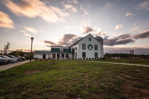 A white farmhouse with a green roof, surrounded by grass and trees, under a cloudy sky at sunset.