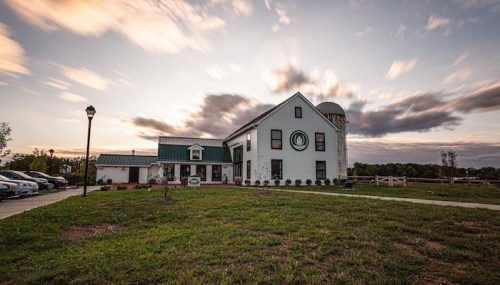A white farmhouse with a green roof and silo, set against a cloudy sunset sky, surrounded by grass and parked cars.