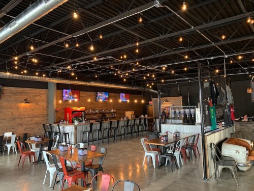 A modern restaurant interior with string lights, a bar, and tables, featuring TVs and a rustic decor style.