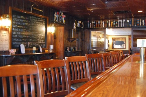 A cozy bar interior with wooden stools, a polished counter, and a chalkboard menu on the wall.
