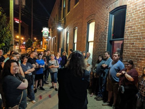 A diverse group of people gathered outside a brick building at night, listening to a speaker.