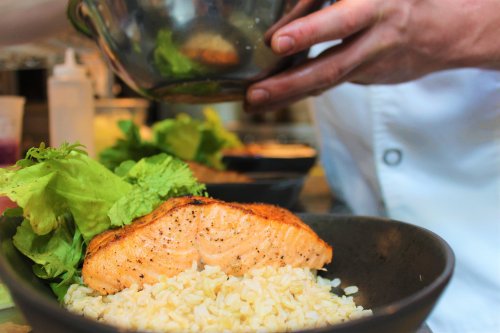 A chef adds seasoning to a plate with salmon, brown rice, and fresh lettuce.