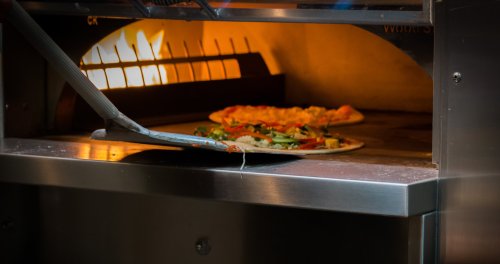 A pizza being pulled from a wood-fired oven, with flames visible in the background.