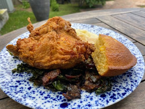 A plate of fried chicken, cornbread, and sautéed greens served on a speckled blue plate outdoors.