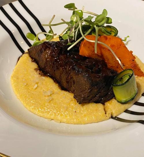 A plated dish featuring tender beef, creamy polenta, and garnished with greens and roasted vegetables.