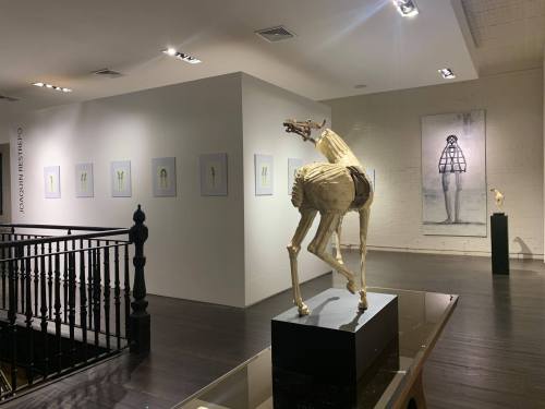 A gallery interior featuring sculptures and framed artworks on white walls, with wooden flooring and modern lighting.