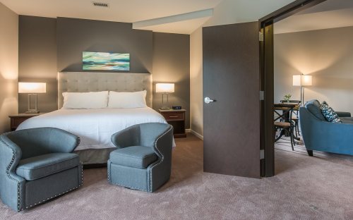 A cozy hotel room featuring a bed, two chairs, and a doorway leading to a small sitting area with a desk.