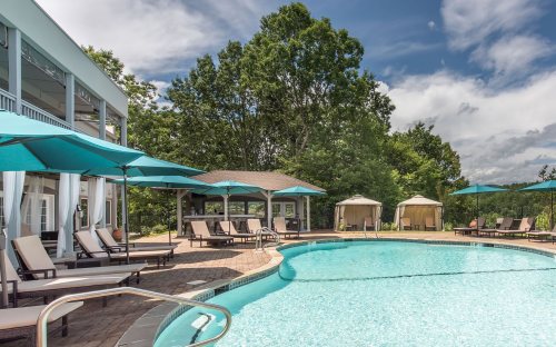 A serene pool area with lounge chairs, teal umbrellas, and cabanas, surrounded by lush greenery and a clear blue sky.