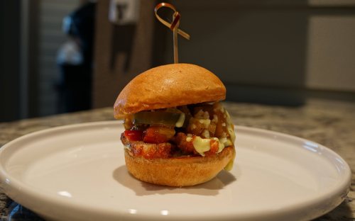 A close-up of a small burger on a white plate, topped with pickles, ketchup, and a toothpick garnish.