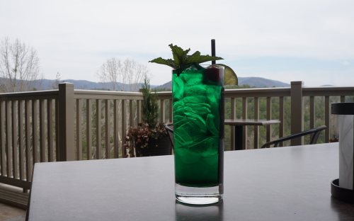 A vibrant green cocktail with mint and a cherry, set on a table with a scenic mountain view in the background.